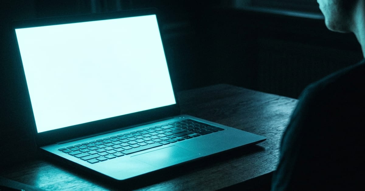 A laptop on a desk in a dark room, its screen a blank white glare with nothing on it