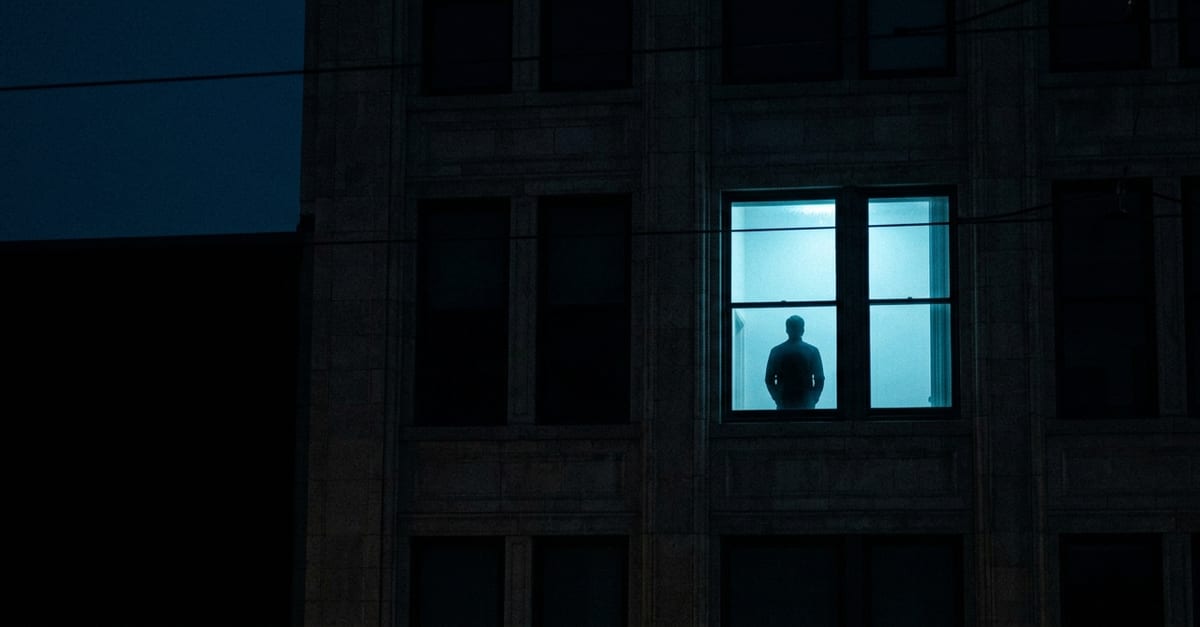 One lit window high in a dark office building at night, every other window black